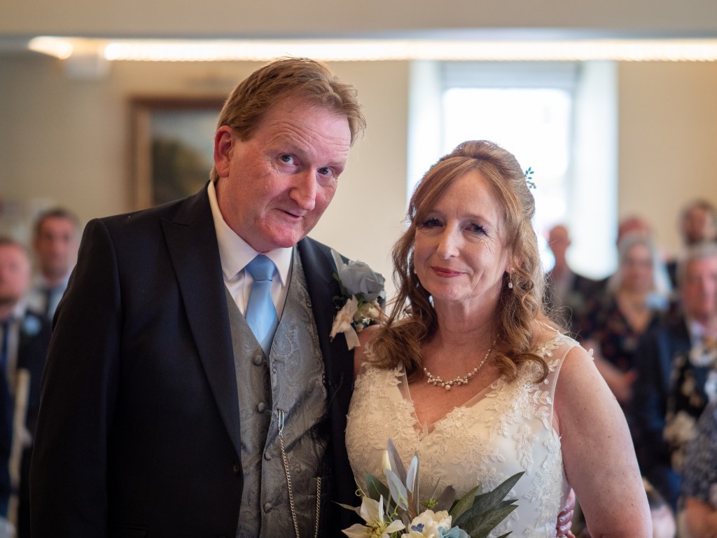 A couple stands together, smiling at the camera, during a wedding ceremony. The man is dressed in a suit and tie, while the woman wears a white wedding dress with floral details. The background features guests and soft lighting.