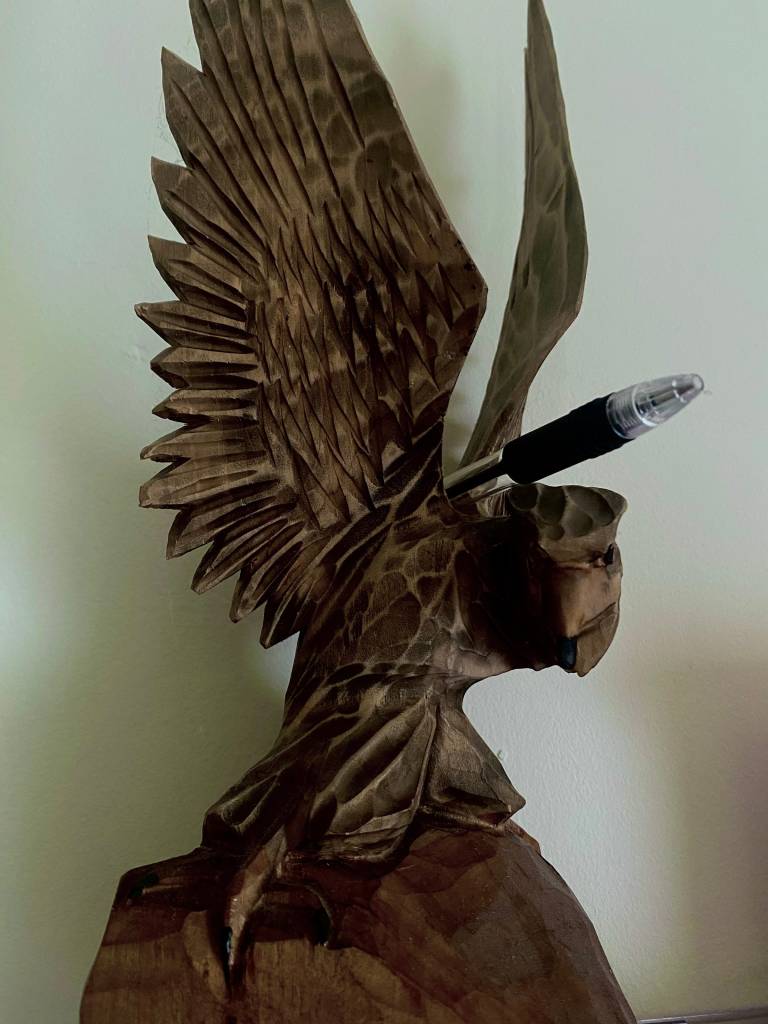 A wooden sculpture of a bird with intricate detailing, featuring a pen inserted in its back, placed against a plain wall.